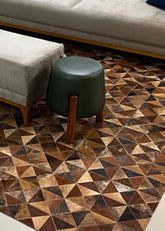 A cowhide patchwork rug featuring a geometric pattern of interlocking triangles in various shades of brown, placed on a floorboard in a living room setting with a couch and stool in the background.